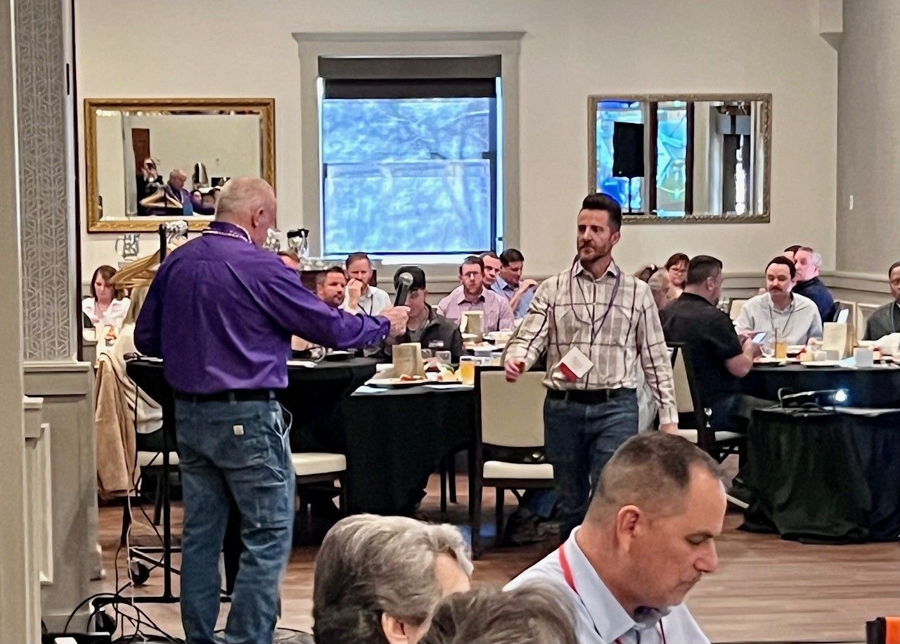 Two men at a business event; one hands a microphone to the other, seated among others at round tables.