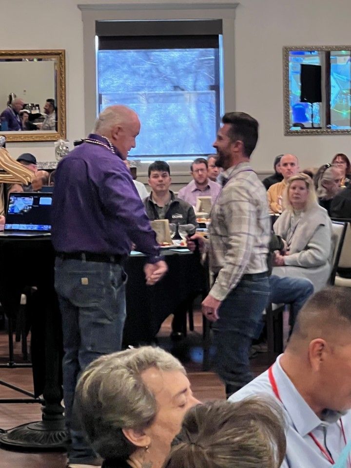 Two men interacting at an event; man in purple shirt shakes hands with man in plaid shirt. Room setting with audience.