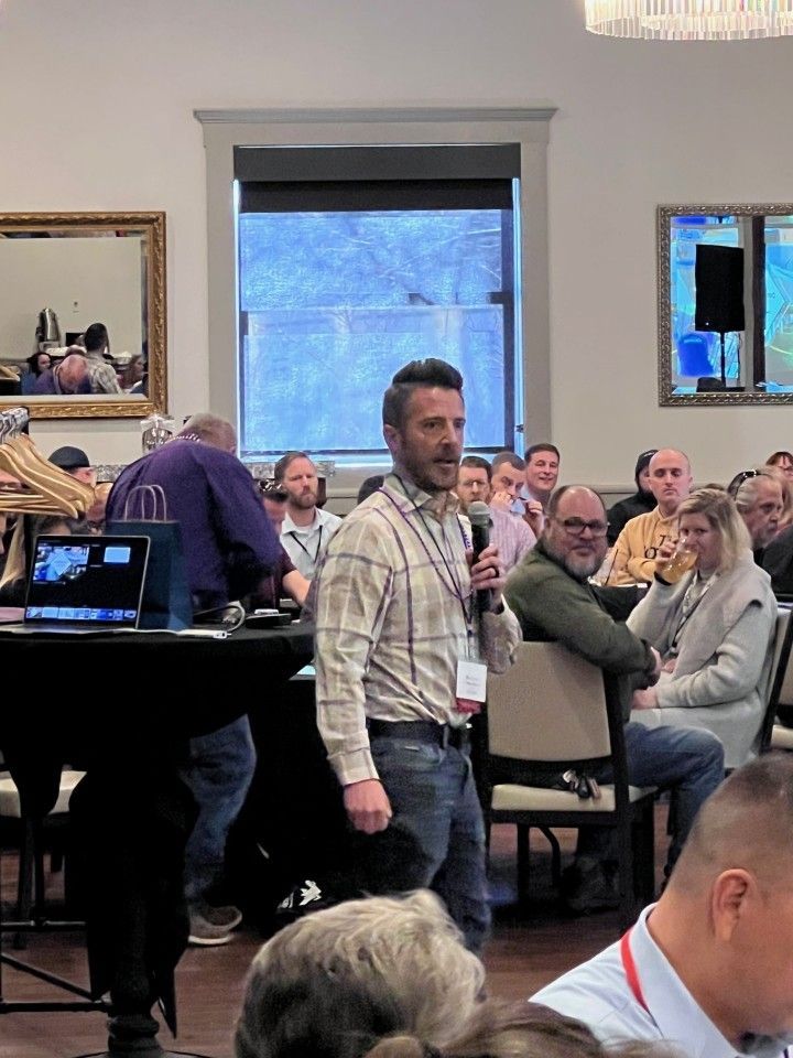 Man speaking into a microphone at an event, facing a seated audience.