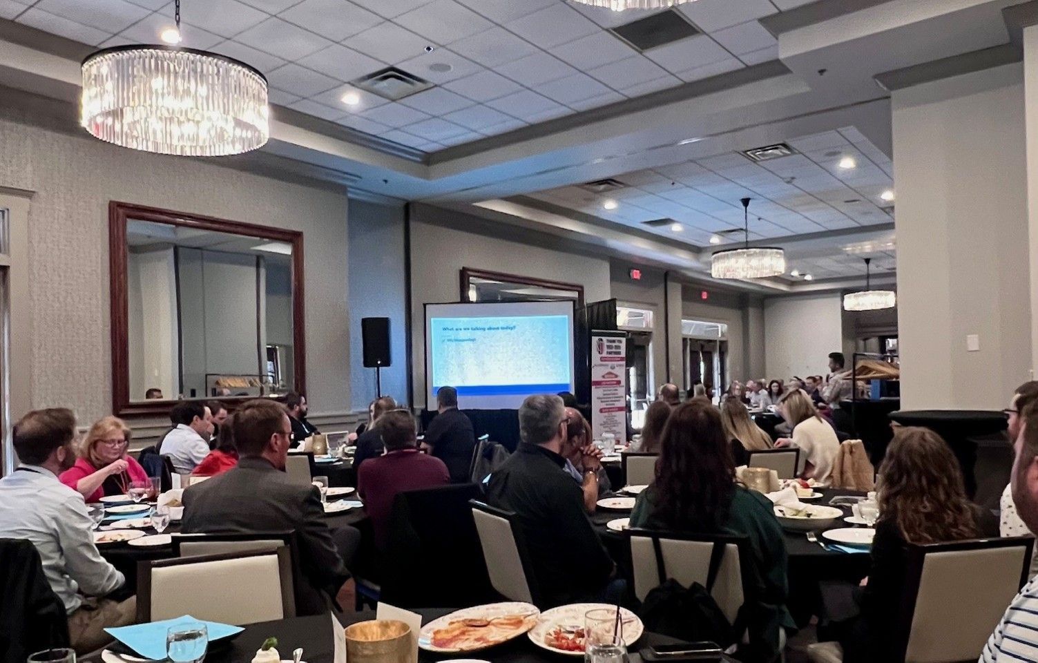 Conference room with attendees seated at tables, a presentation on screen, and overhead chandeliers.