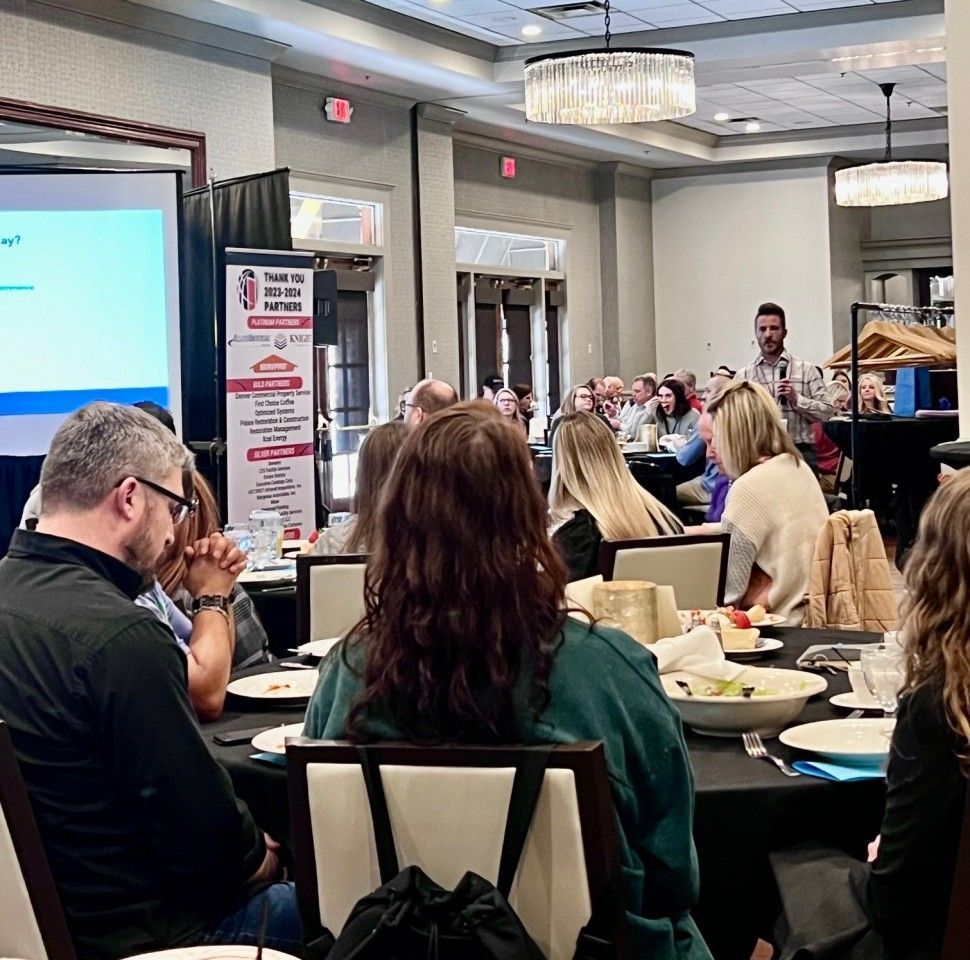 People attending a business presentation in a large hall; a man speaks, a screen displays information.