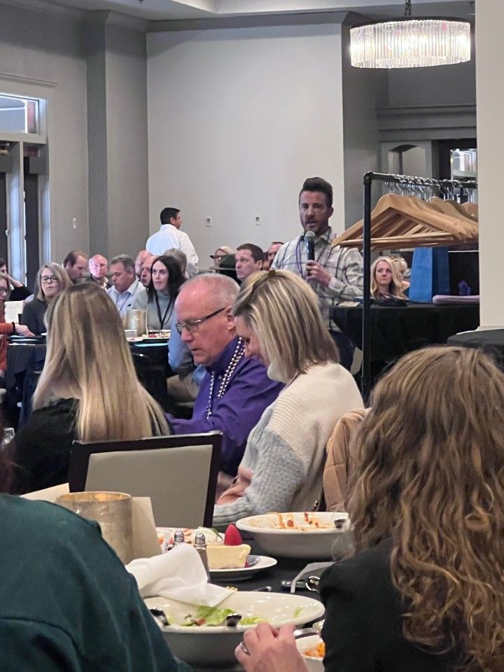 Man speaking into a microphone at a conference. People seated at tables listen in a hotel ballroom.