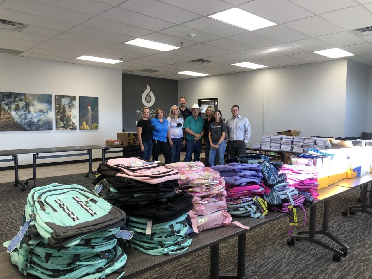 People gather around tables with stacked backpacks. Boxes and artwork are also present in a well-lit room.