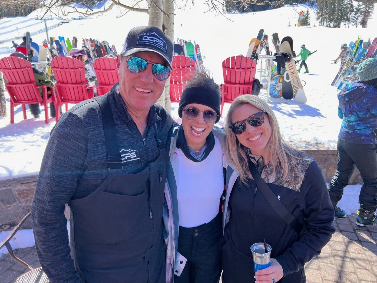 Three people smiling for a photo outdoors at a ski resort, with snow and red chairs in the background.
