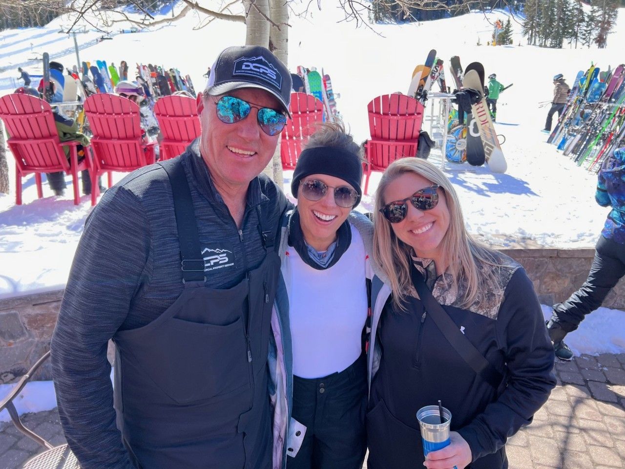 Three people smiling outside in the snow. Red chairs and skis in background.