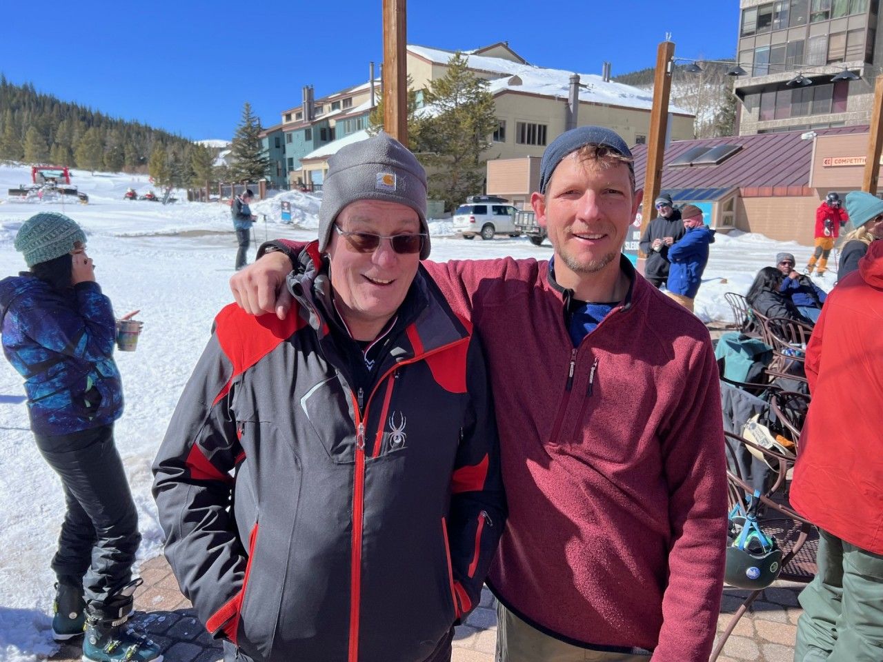 Two men pose outdoors in the snow, one with arm around the other. Buildings and skiers in the background.