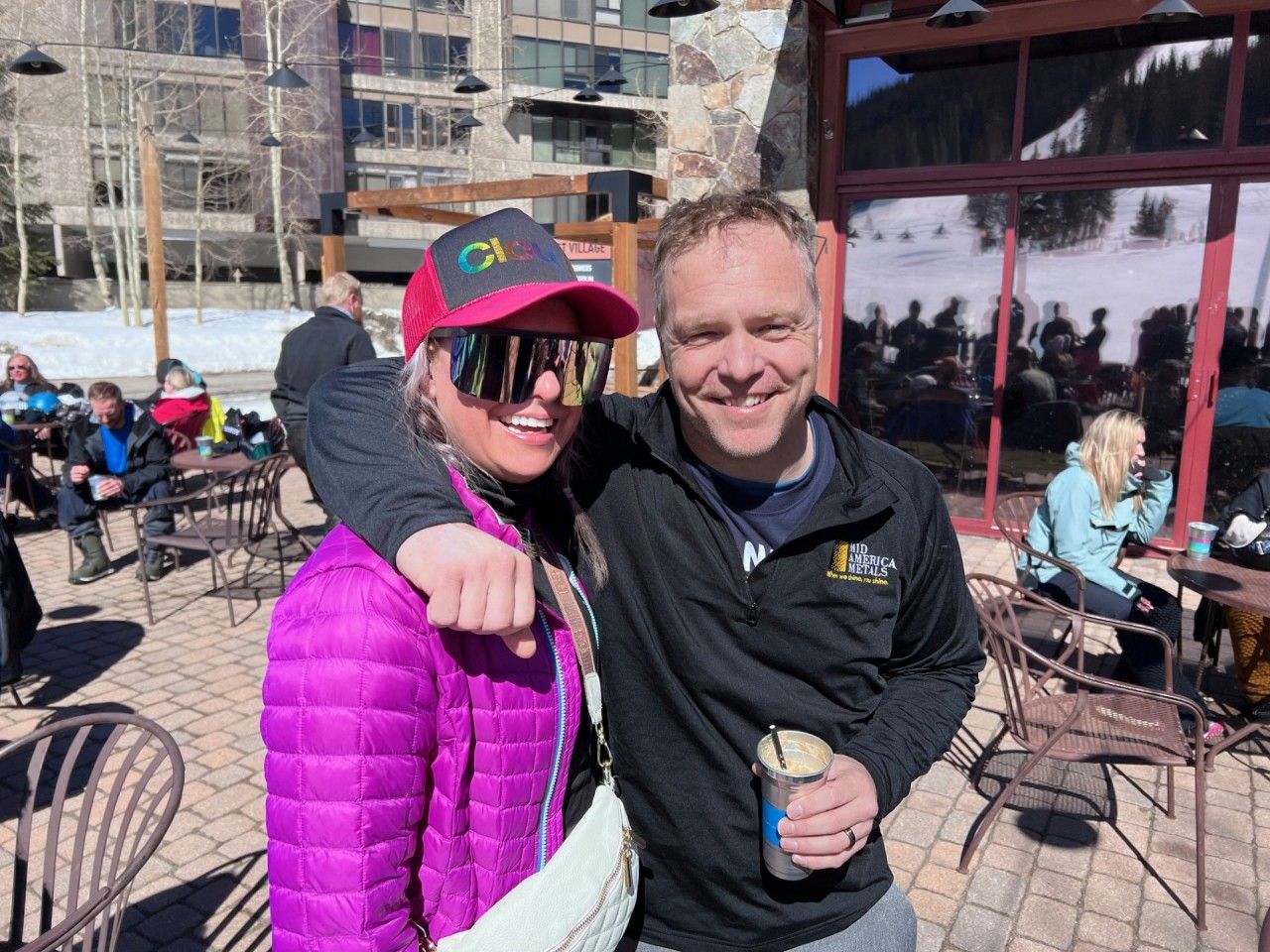 Two people smiling, posing outdoors. Woman in pink jacket and sunglasses, man in black jacket holding a drink.