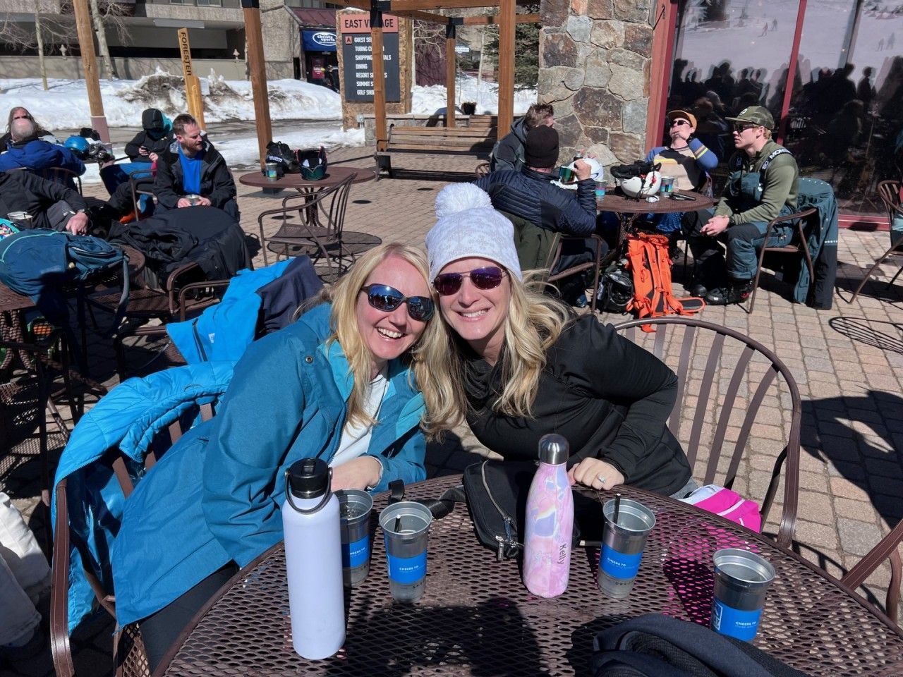 Two women smile outdoors at a cafe table, with water bottles, other patrons, and a snowy mountain backdrop.