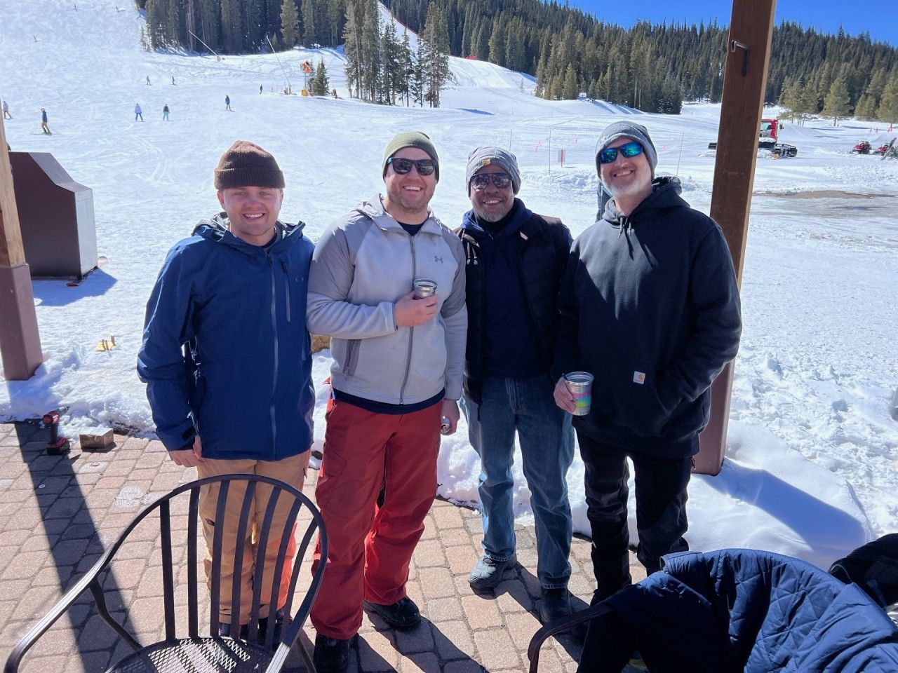 Four people stand in front of a snowy ski slope on a sunny day.