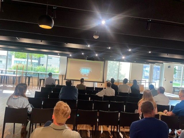 Audience seated in a room, watching a presentation on a screen. Bright light and window in the background.