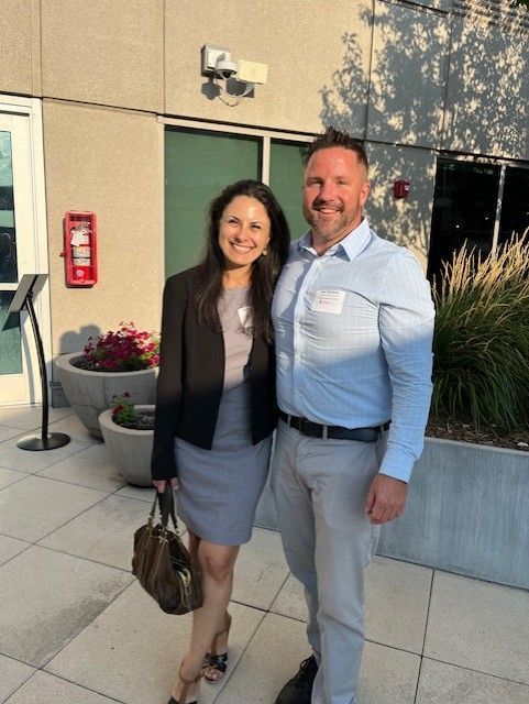 A woman and a man pose together outdoors in front of a building. They are both smiling.
