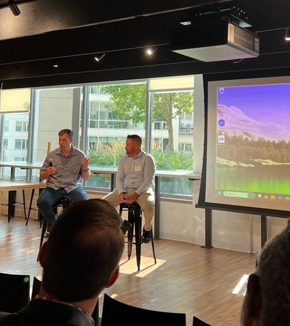 Two men on stools at a panel, speaking in a room. Projector shows a mountain scene.
