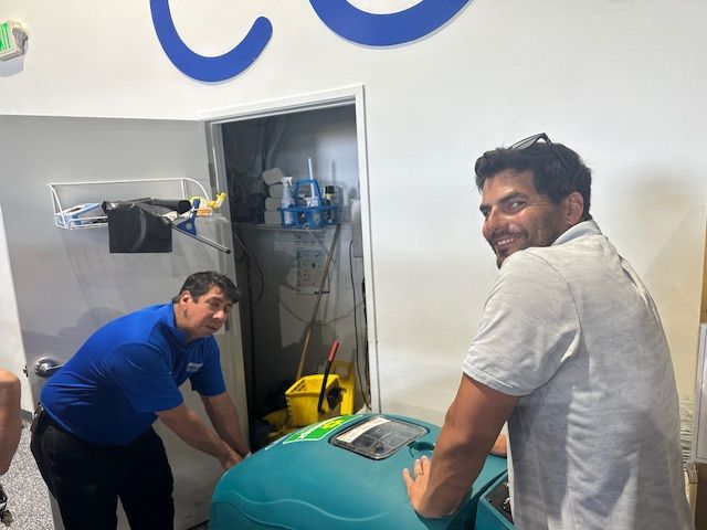 Two men moving a teal cleaning machine into a utility closet; one man smiles.