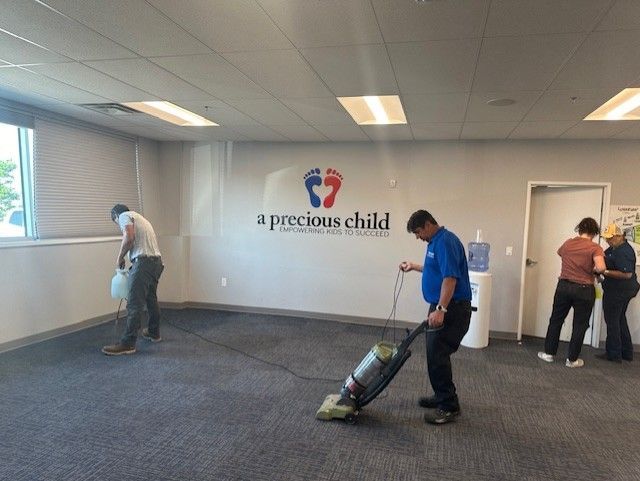 People cleaning an office. One vacuums carpet, another empties a trash bag, and two stand by a doorway.