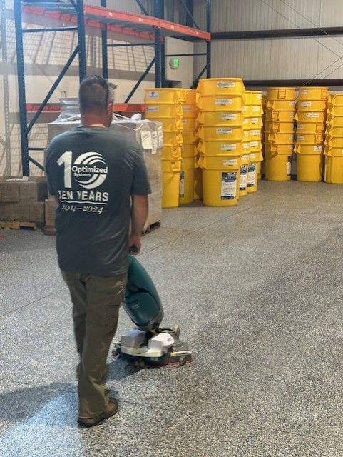 A person using a floor buffer in a warehouse, with stacks of yellow buckets in the background.