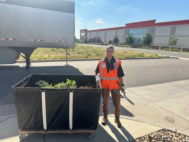 Person in orange vest stands by a large bin of yard waste; a semi-truck and building are in the background.