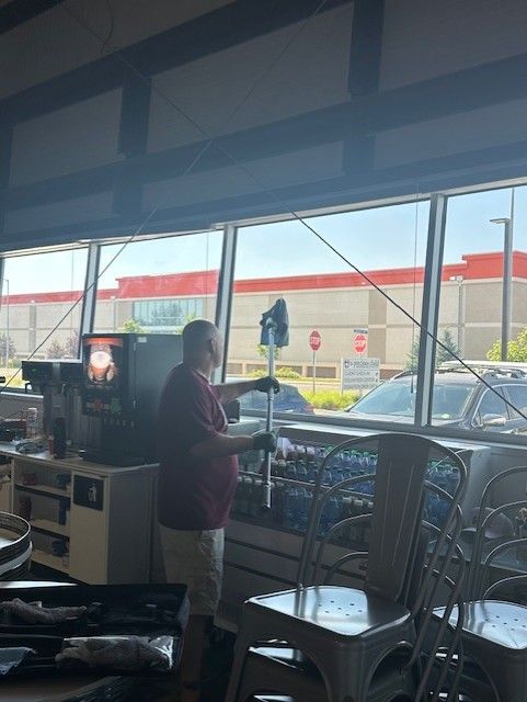 Man cleaning windows inside a store, using a long-handled tool. Behind him, a row of windows and an exterior view.