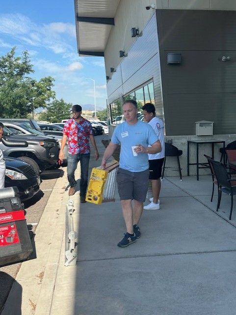 Man carrying a toolbox and coffee walks by a building with two others. Sunny outdoor setting.