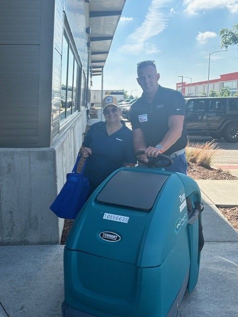 Two people by a teal floor cleaner outside a building, sunny day. One holds a broom.