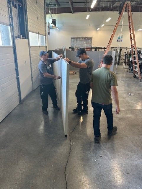 Three people installing a wall in a large, bright warehouse. Gray wall panels, concrete floor.