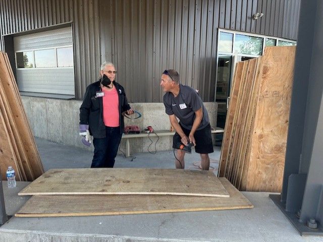 Two people cutting plywood outdoors. One holds a power tool, the other looks on. Building in background.