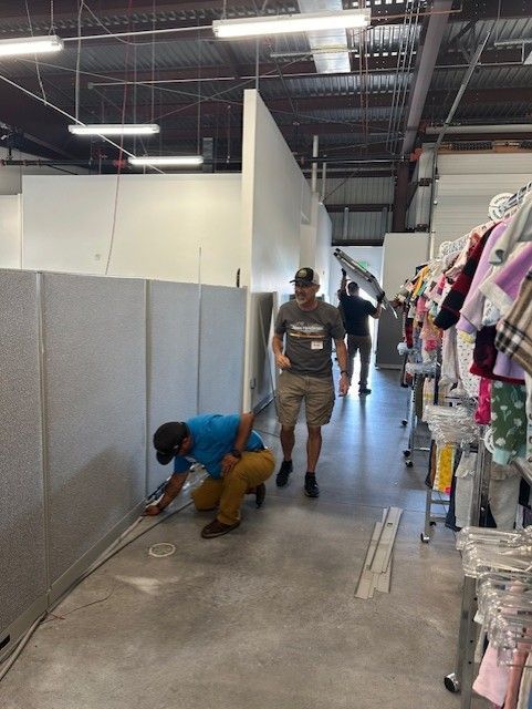 Men working inside a retail space. One kneels, working on wiring. Others walk by racks of children's clothing.