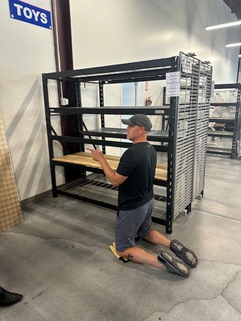 Person kneeling, working on black shelving unit in a store, holding what appears to be a cell phone.