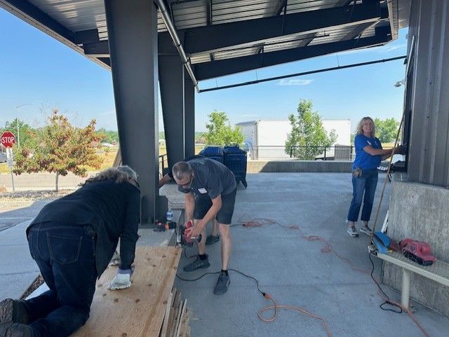 Three people working outdoors under a shelter: one kneeling, sawing; one using a drill; and one working on a wall.