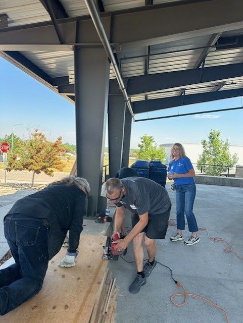 People working on a construction project: one using a saw, another kneeling, and a woman watching. Outdoor setting.