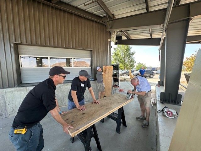 Four men building with wood outdoors near a building with a garage door. One cuts wood with a saw.
