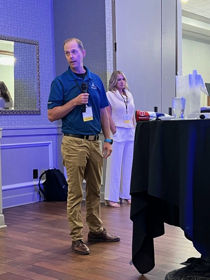 Man speaking into a microphone at an event, another person standing behind him, near a table.