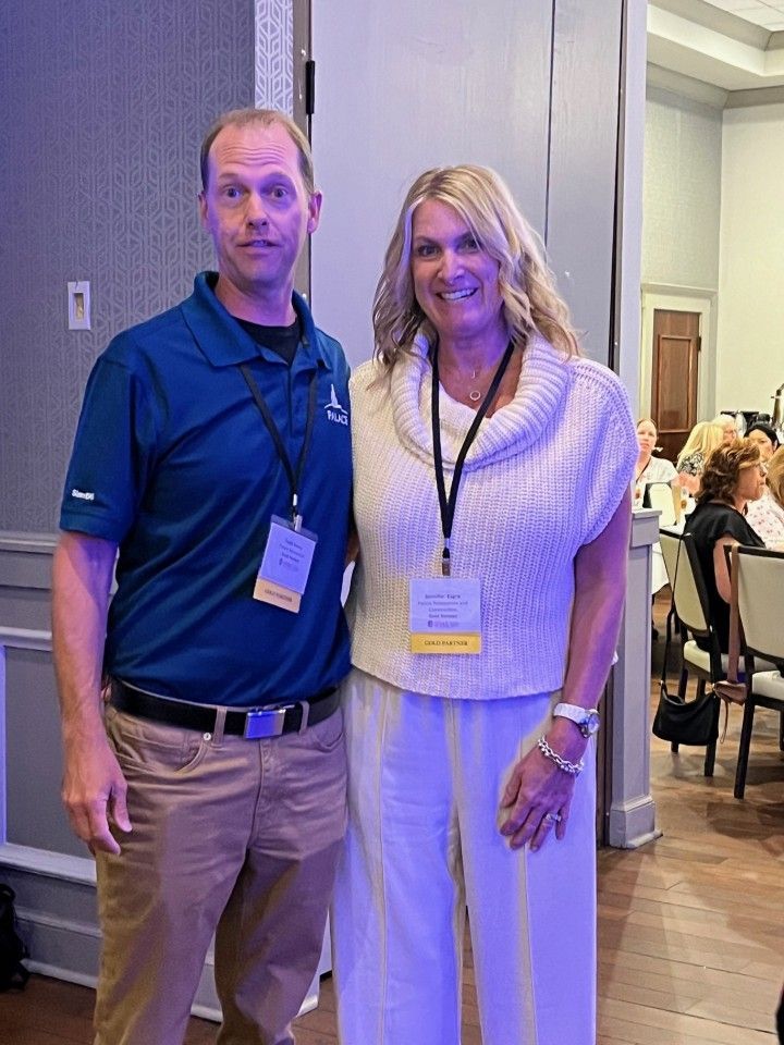 Man and woman standing together indoors, both wearing lanyards with name tags, smiling slightly.