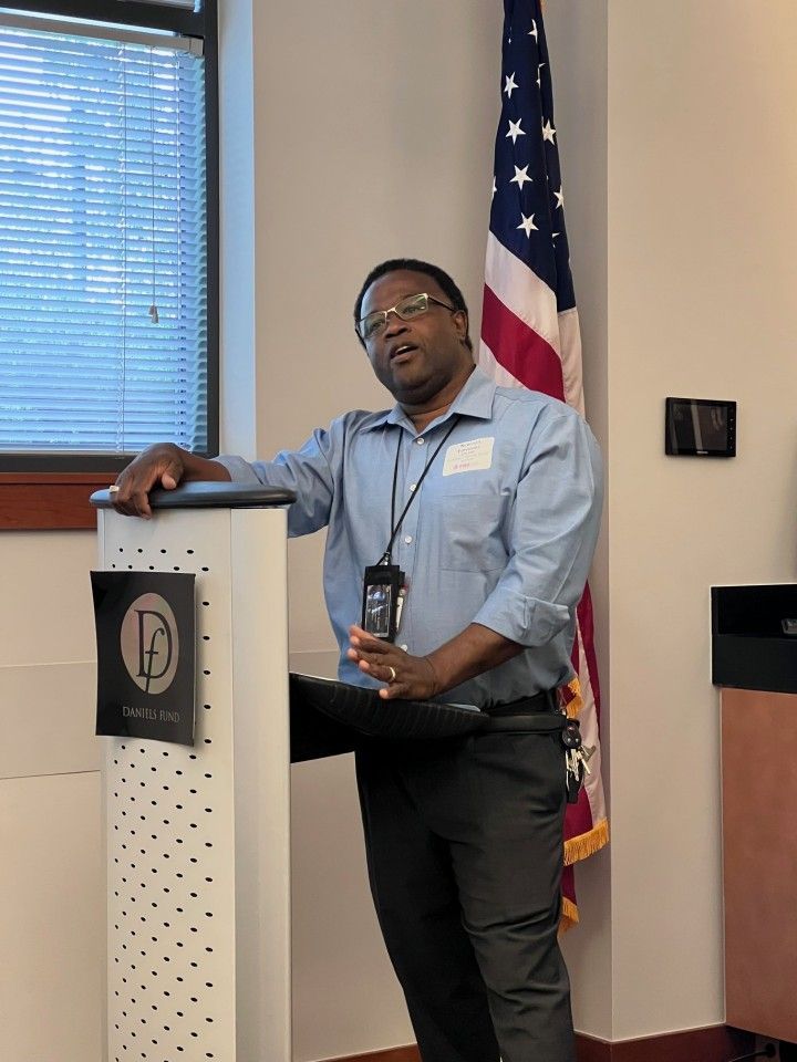 Man speaking at a podium, American flag in the background. He wears a light blue shirt and dark pants.