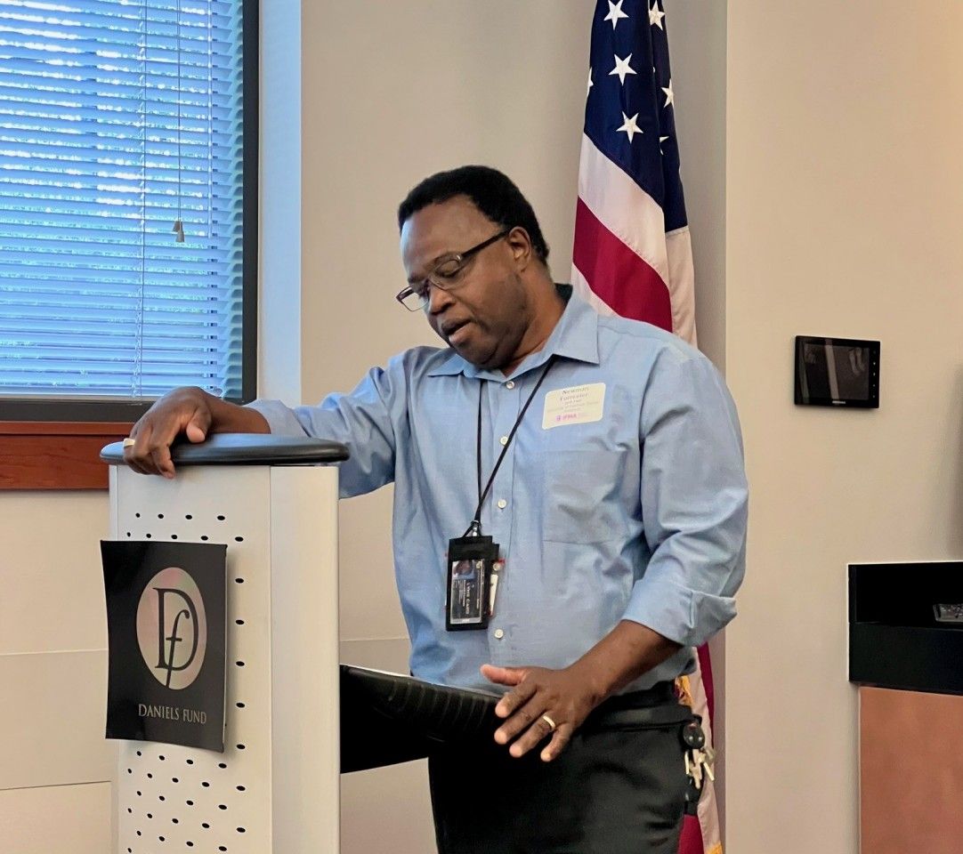 Man leaning on a podium, speaking, indoors near an American flag.