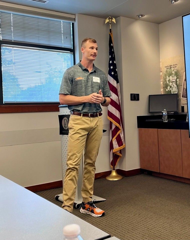 Man giving a presentation, standing near American flag. He's wearing a green shirt and khaki pants. Indoor setting.