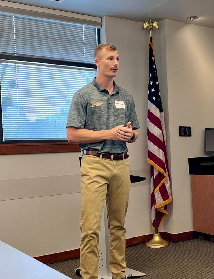 Man speaking, hands clasped, in front of a U.S. flag. He wears a green polo, tan pants, and stands in a room.