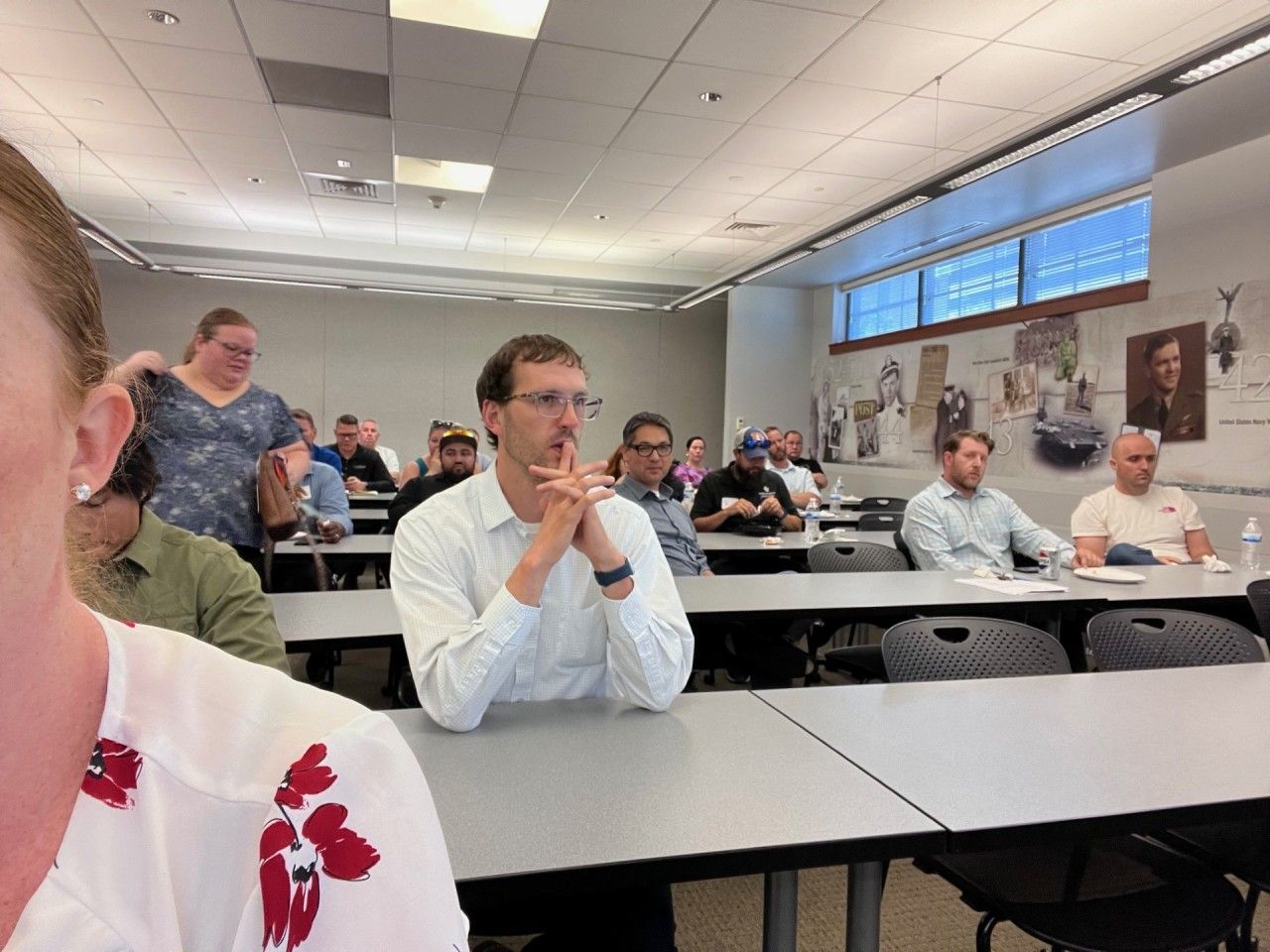 People seated at tables in a classroom setting. A person takes a selfie, visible in the foreground.