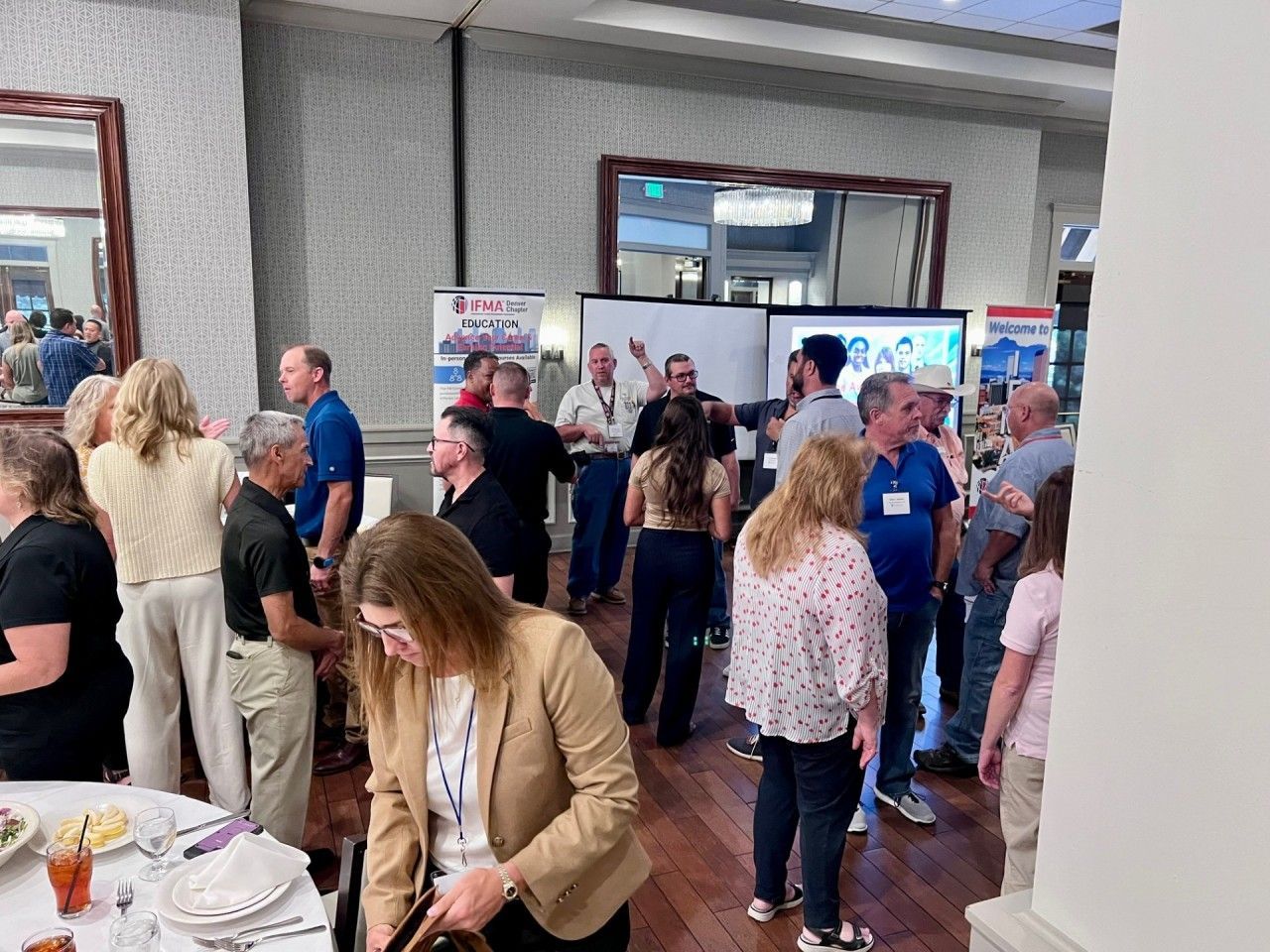 Group of people at an event in a well-lit room; some are gathered near tables, others by display boards.
