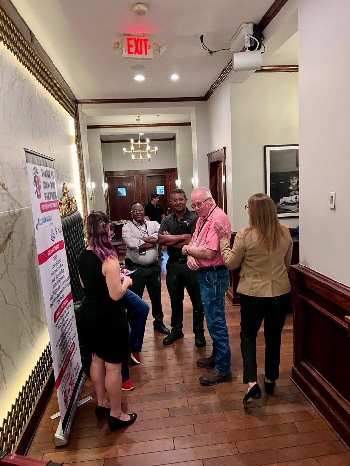 People conversing in a hallway with a banner. Exit sign overhead, wood floor.