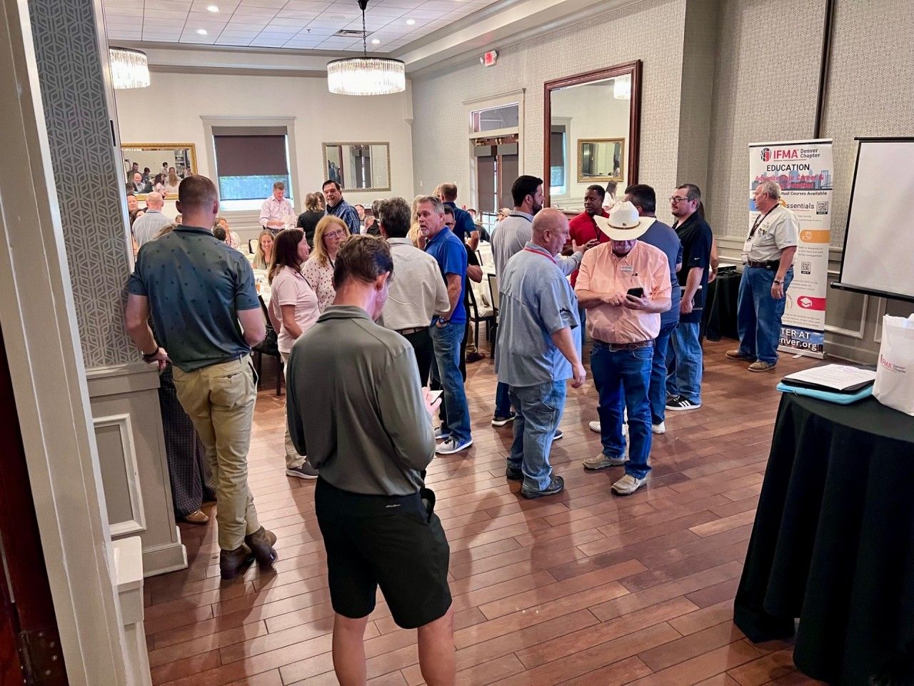 A group of people standing in a room, some looking at phones, others talking. Conference or event setting.