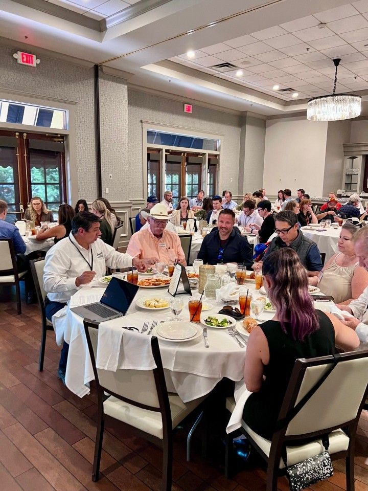 People seated at round tables in a restaurant, eating and conversing. Bright dining room with white tablecloths.