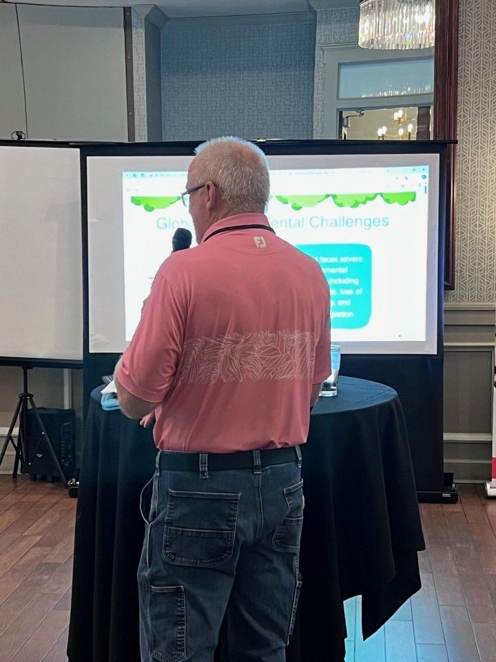Man in pink shirt giving a presentation, standing in front of a screen with text, at a table.