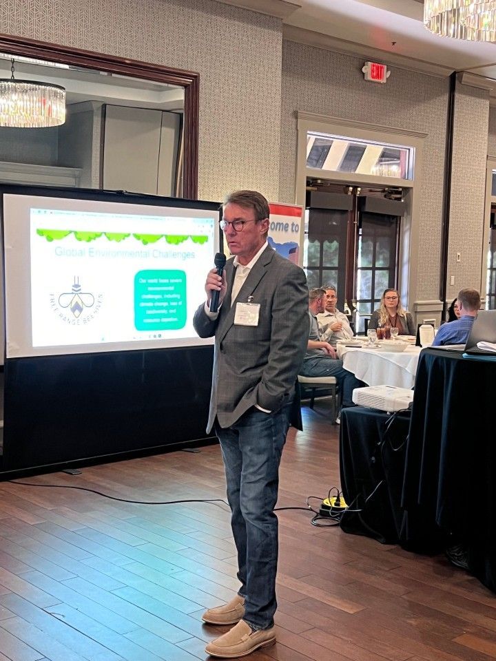 Man in blazer speaks at a presentation, using a microphone. Audience in background.
