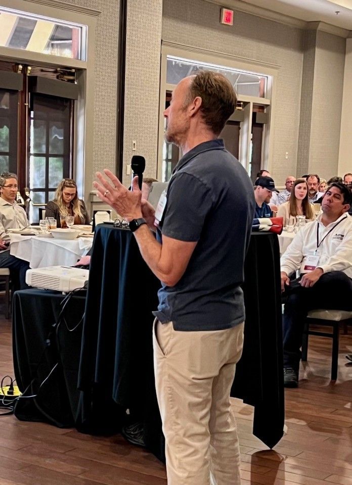 Man speaking at an event, gesturing with hands. Standing at a podium, with an audience seated at round tables.