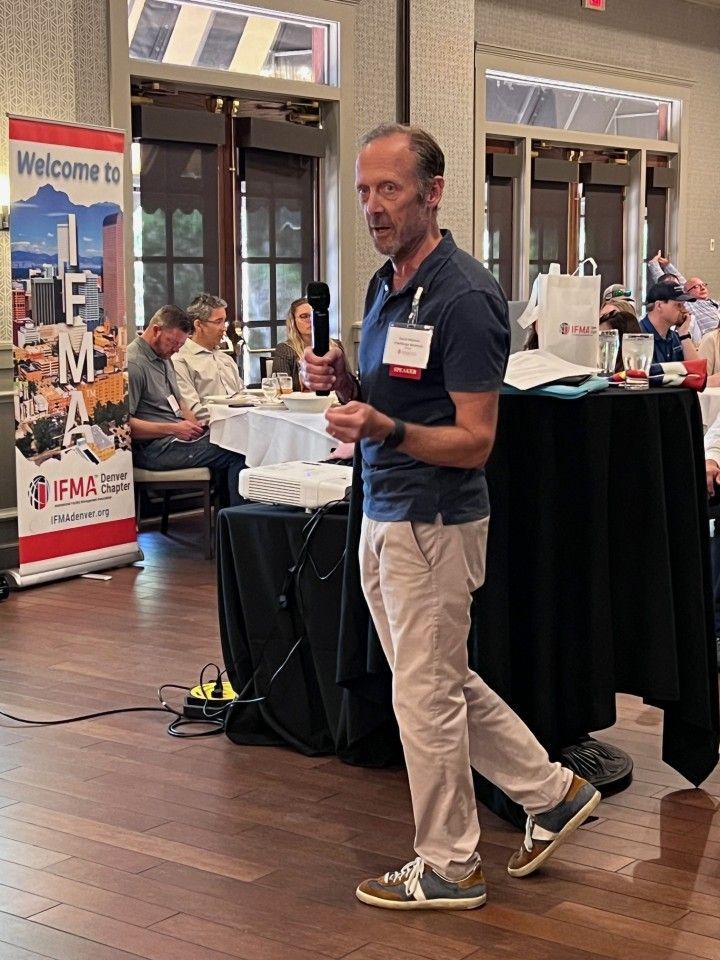 Man speaking at a conference, holding a microphone. Indoor setting with a banner and tables.