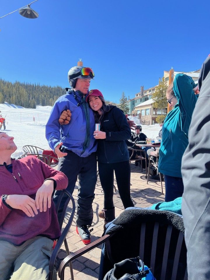 People embrace outdoors in the snow. Man in blue jacket, woman in black hugging. Sunlit, outdoor café setting.