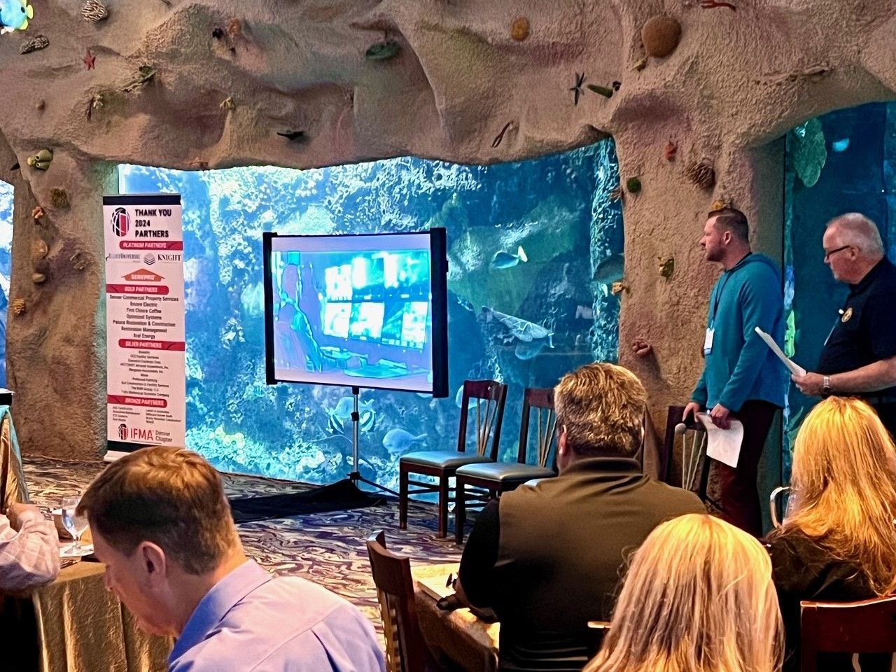 People at a presentation in front of a large aquarium tank. Two men speak near a screen and a poster.
