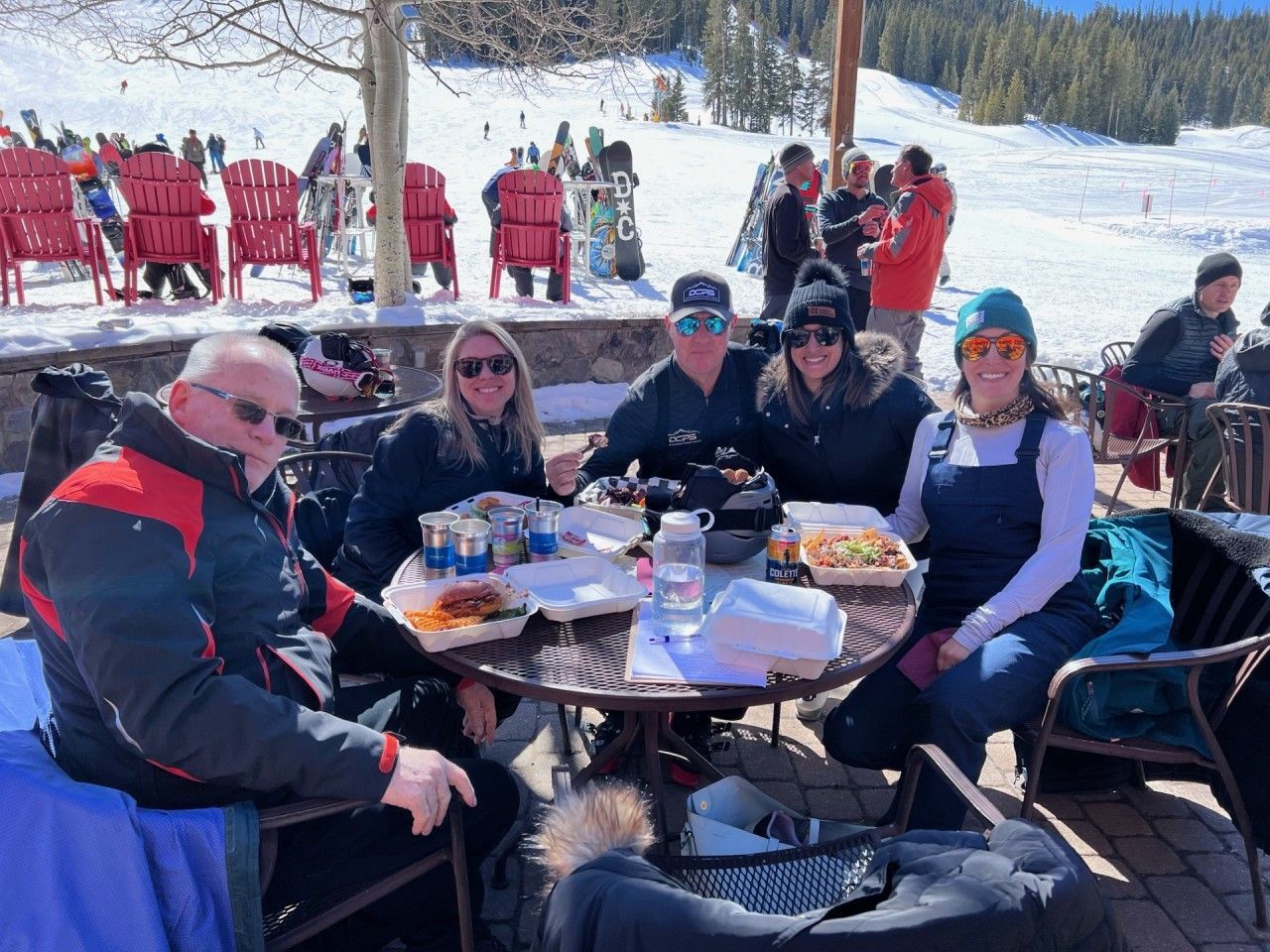 Five people seated around a table, eating lunch outdoors on a sunny, snowy ski resort deck.