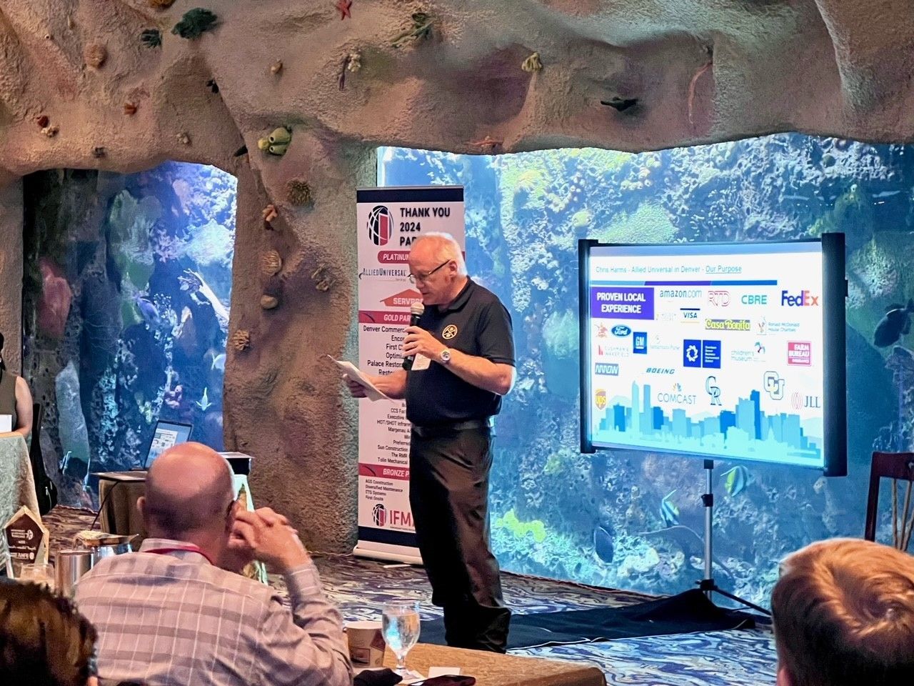 Man presenting at a conference. Audience in foreground, screen with logos, backdrop is an aquarium.