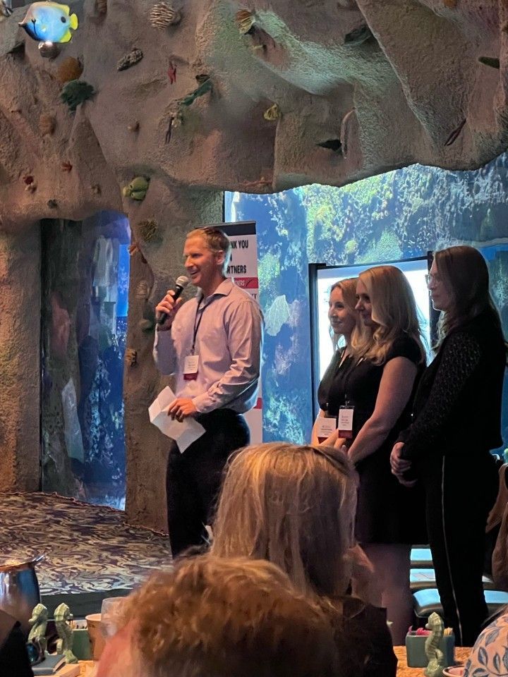 Man speaking at a podium, three women standing behind him. Aquarium backdrop, event setting.